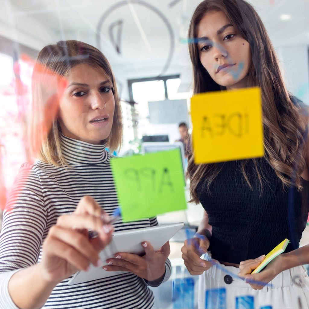 Shot of two business young women working together on wall glass with post it stickers in the modern startup office.